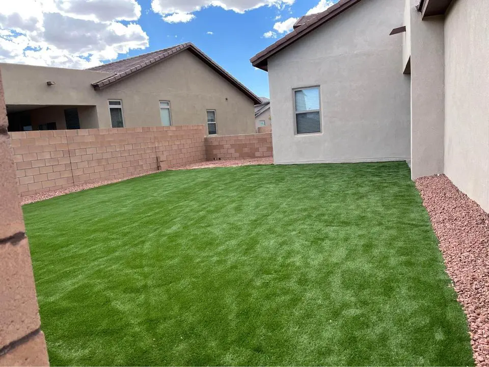 A backyard with neatly manicured green grass, courtesy of Casa Grande Turf & Paver, is surrounded by beige stone and brick walls. Two adjacent beige houses frame the scene under a partly cloudy blue sky. Small rock beds border the lush lawn along the walls.