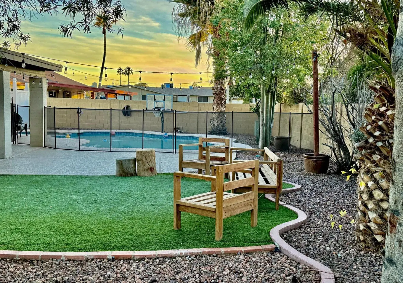 The backyard, expertly designed by Casa Grande Turf & Paver, features a swimming pool encircled by a safety fence. An artificial grass area is fitted with wooden benches beneath string lights, while a vibrant sunset paints the sky above various trees.