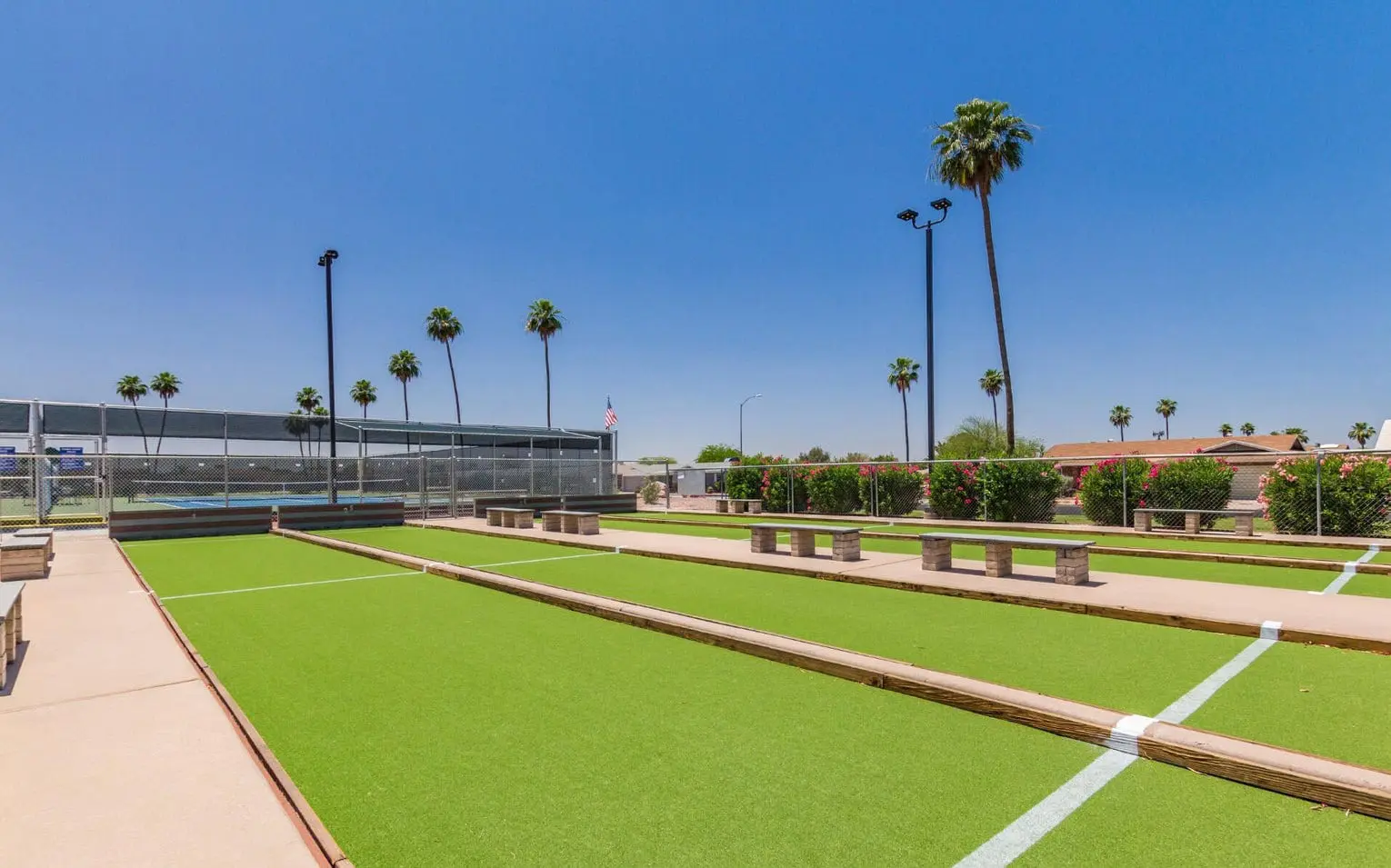 A sunny outdoor scene showcases a bocce ball court beneath a bright blue sky, with palm trees swaying in the background and lush green turf expertly laid by Casa Grande Turf & Paver. Benches line the court's sides, and a fenced area adds charm to this inviting space.