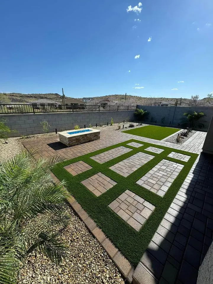 A backyard with neatly divided green grass sections and brick pathways. A stone fire pit is in the center, surrounded by desert landscaping. A block wall and distant desert scenery are visible under a clear blue sky.