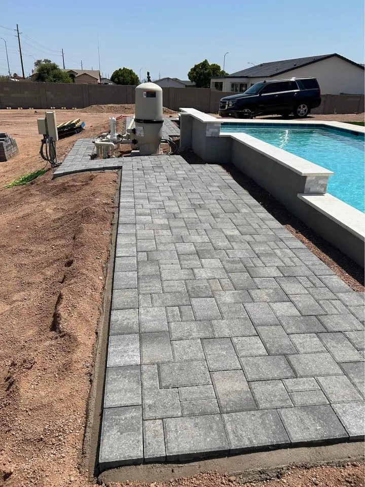A newly constructed swimming pool with a gray stone paver walkway beside it. A black SUV is parked on the soil in the background, next to some construction materials and equipment. The area appears to be in a residential neighborhood.