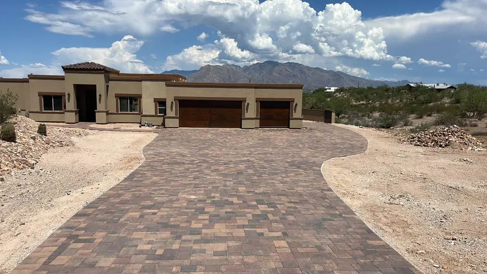 A modern, single-story desert home with light-colored walls and a dark wooden garage door. The house is surrounded by a rocky landscape, and a long, patterned brick driveway leads to the entrance. In the background, mountains and a cloudy sky are visible.