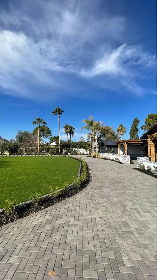 A paved walkway curves through a vibrant green lawn lined with palm trees under a clear blue sky. On the right, modern houses with wooden accents are visible. The scene is peaceful and well-maintained, with lush greenery.