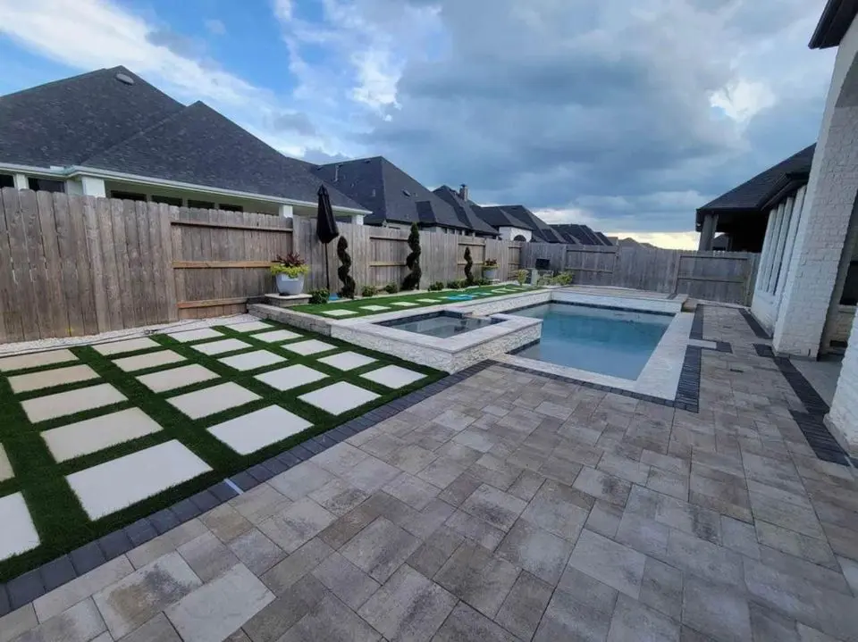 A backyard features a swimming pool with an attached hot tub. The area is surrounded by stone tiles and decorative grass patterns. There's a wooden fence and houses in the background under a partially cloudy sky.