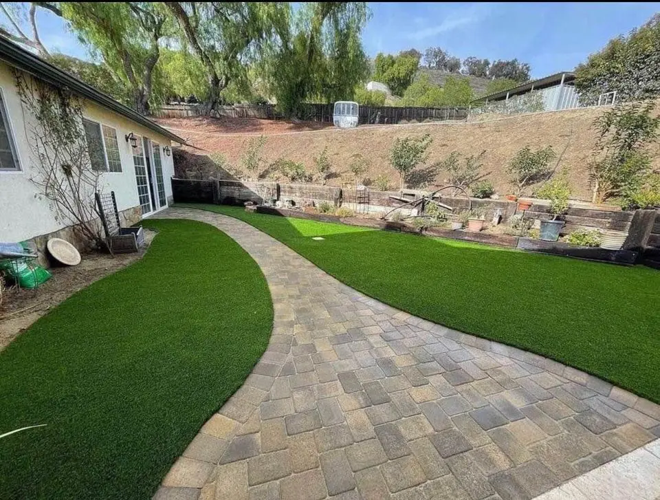 A backyard with a stone path curving between areas of artificial grass. The area is surrounded by a hillside with sparse greenery, and is adjacent to a house with a white exterior. There are planters and garden items along the path.