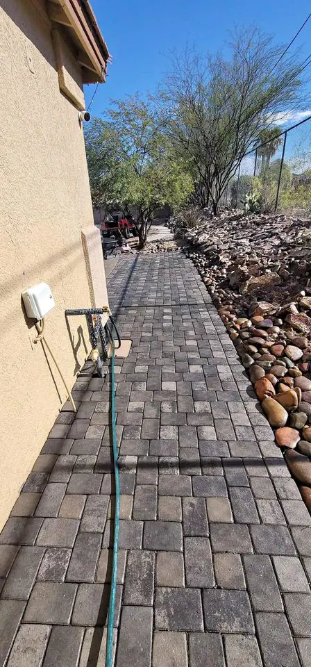 A paved walkway beside a house leads through a desert landscape with scattered stones and sparse trees. A garden hose lies along the path, and there's a clear blue sky overhead.