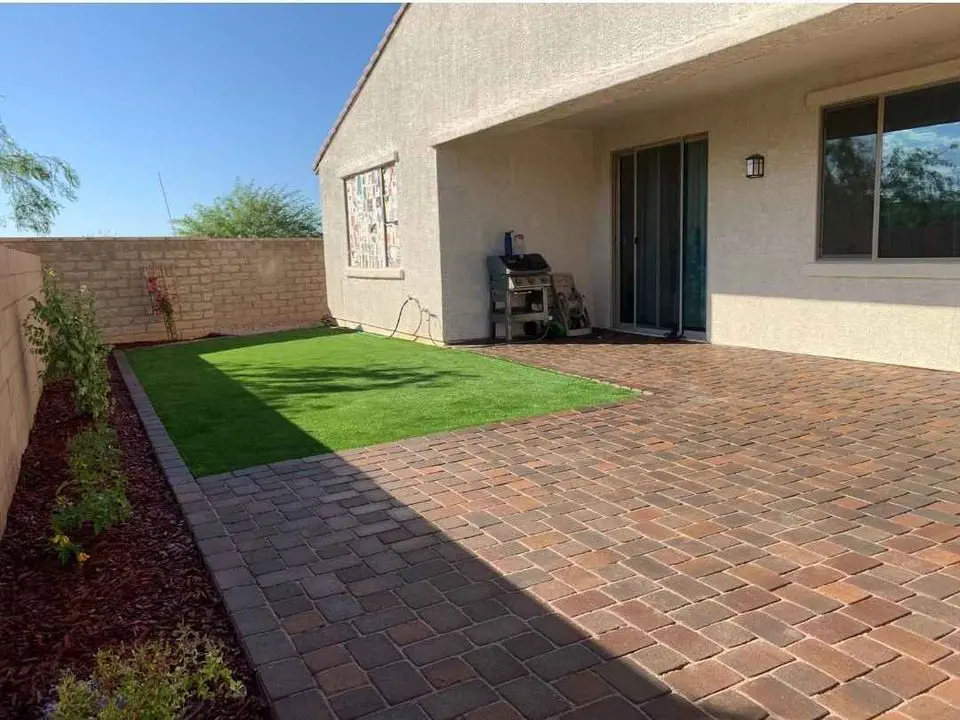 A backyard with a paved patio and a small patch of artificial grass. There is a barbecue grill near the sliding glass door, and flower beds line the left side. A brick wall encloses the area with a view of trees in the background.