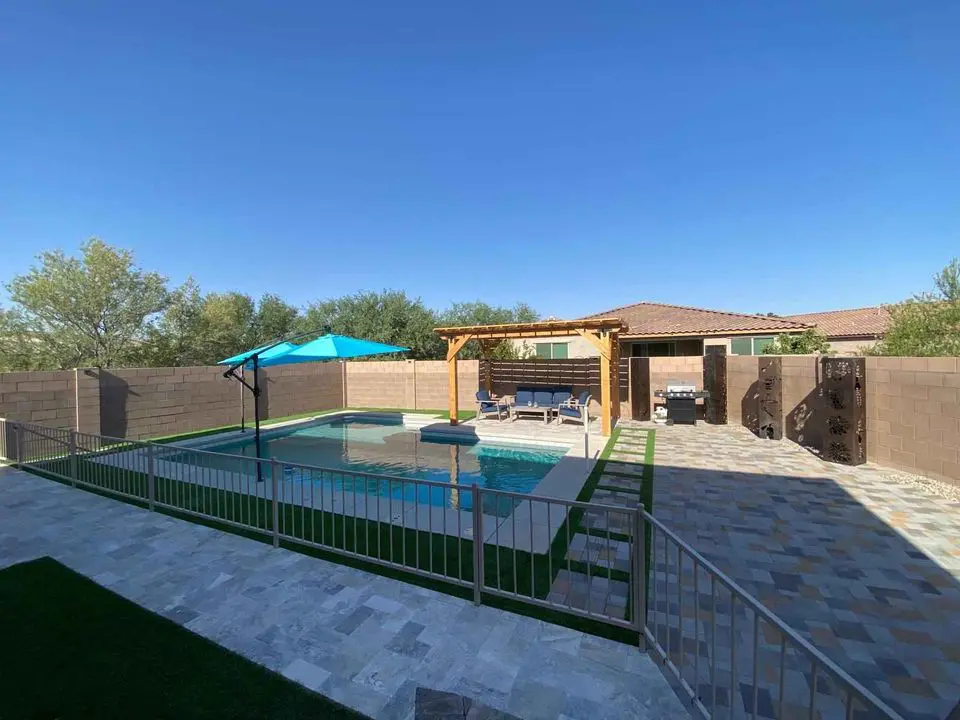 A backyard features a swimming pool with a turquoise umbrella and a pergola-covered seating area. The space is surrounded by a stone patio and a low fence, with trees visible over a privacy wall.