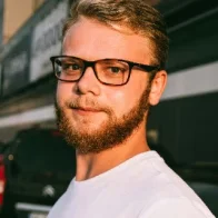 A man with glasses and a beard, wearing a white T-shirt, stands outdoors near a Casa Grande Turf & Paver site. The partial view of a building and parked car in the background is illuminated by the late afternoon sun.