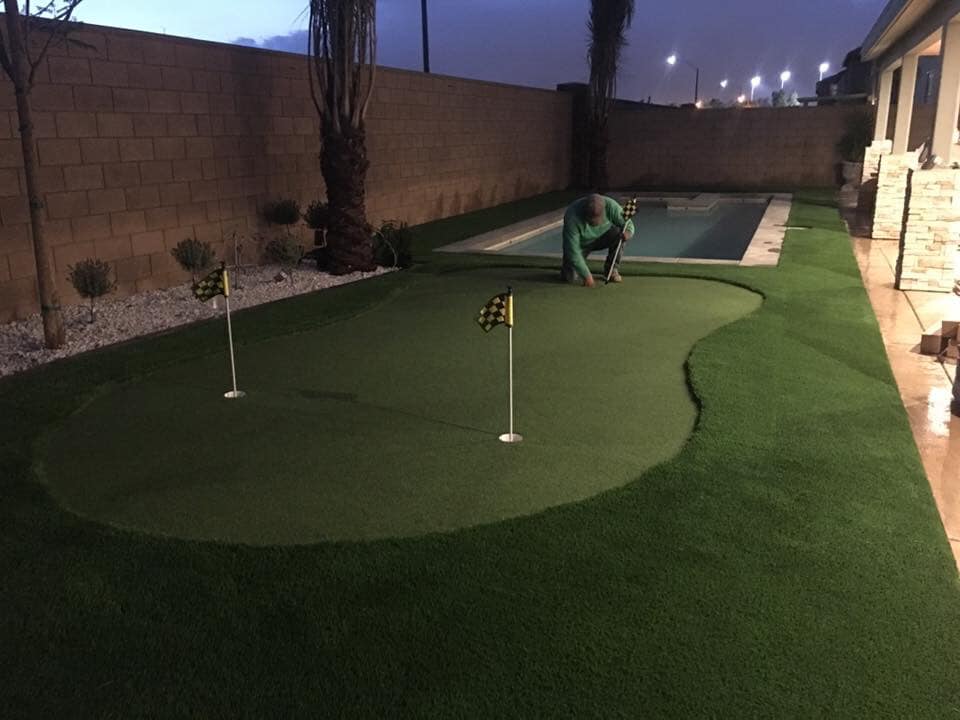 Setting up a mini-golf course in the backyard at night, the scene features artificial grass resembling professional backyard putting greens. With two small golf flags gently swaying, warm lights illuminate Casa Grande AZ's serene evening setting, while a swimming pool shimmers on the side.