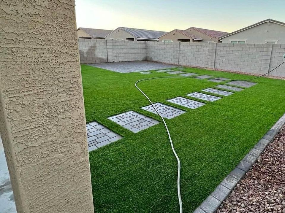 A backyard with a neatly manicured lawn featuring Casa Grande Turf & Paver square stones arranged in a path. A hose lies across the grass, framed by a light-colored brick wall, with neighboring houses visible in the background.