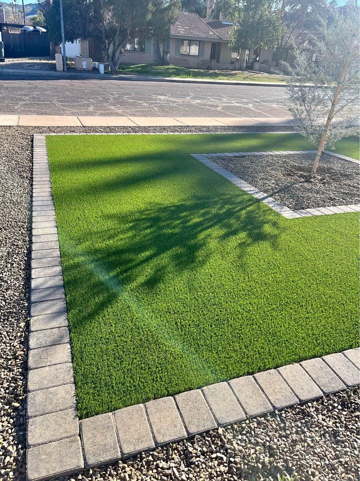 Experience a beautifully maintained front yard featuring a rectangular patch of green artificial grass, expertly installed by Casa Grande Turf & Paver. Light-colored bricks border the lawn, with gravel surrounding it and a tree in the corner. Residential streets and houses complete the serene setting.
