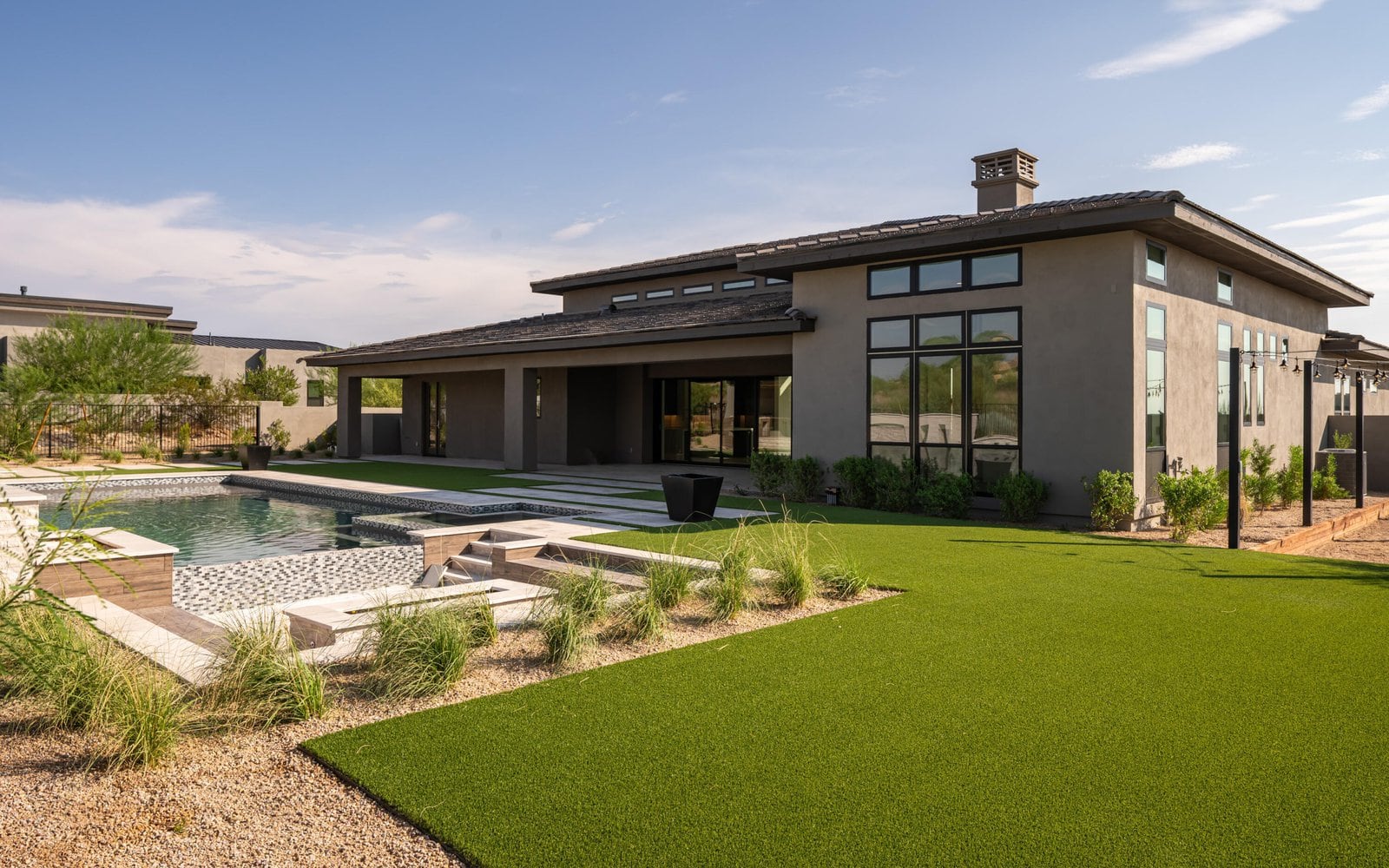 Modern single-story house with large windows and a flat roof, featuring a backyard with a swimming pool surrounded by Casa Grande Turf & Paver's artificial grass and minimalist landscaping under a clear blue sky.