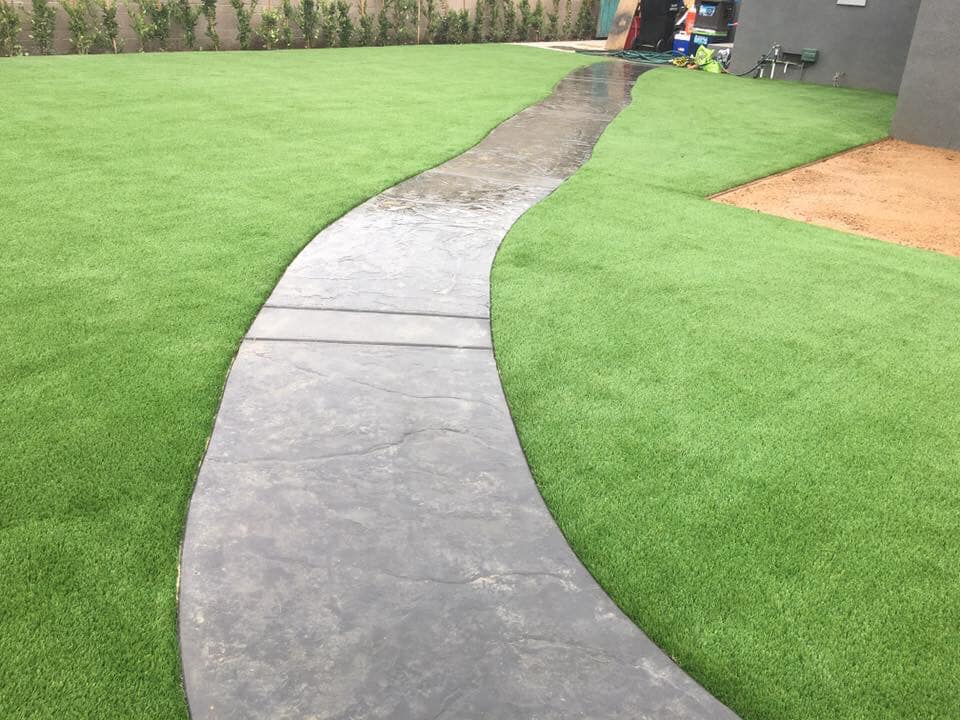 A winding concrete pathway gleams with rain, flanked by vibrant green artificial grass from Casa Grande Turf & Paver. A beige patch of soil appears in the upper right, while a dark gray building peeks through the misty backdrop, capturing the artistry of an expert artificial grass installer in Casa Grande, AZ.