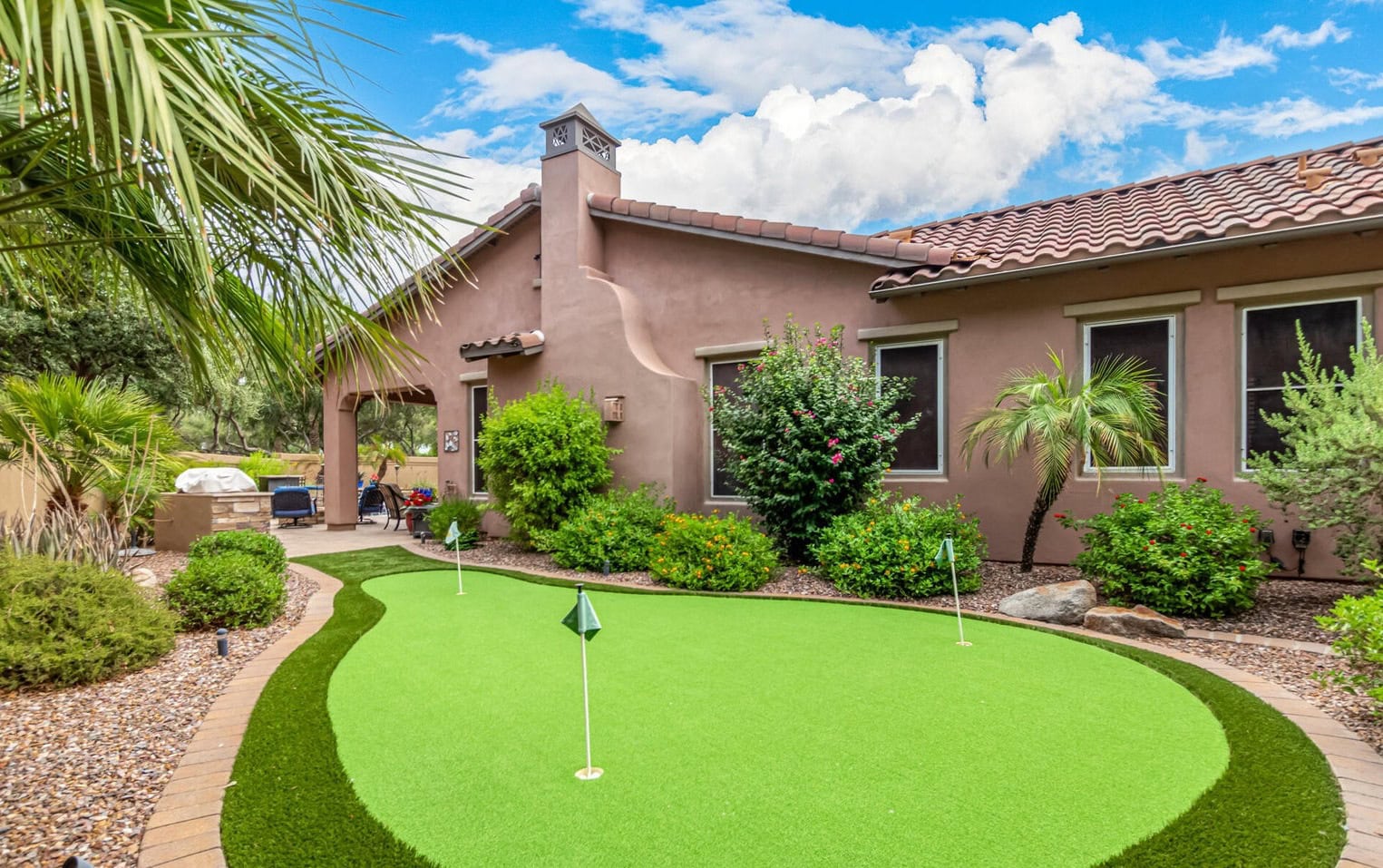 A backyard putting green bordered by stone pathways and lush shrubs adds charm to this Casa Grande, AZ oasis. The brown stucco house with a red-tiled roof is complemented by palm trees and plants, all under a bright blue sky with fluffy clouds.