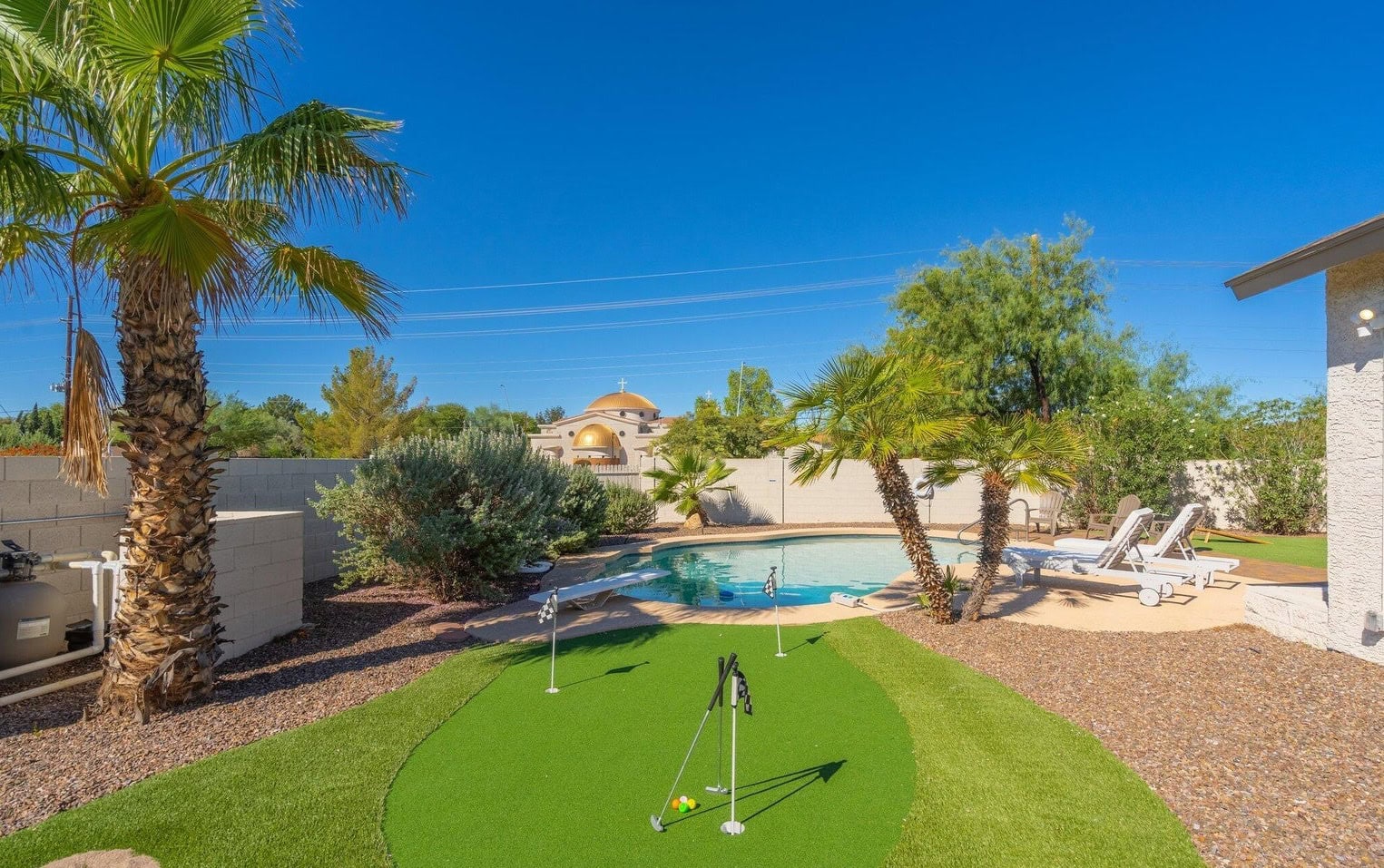 A backyard oasis in Casa Grande, AZ, showcases a lush putting green surrounded by palm trees and plants. In the background, a curved swimming pool with lounge chairs sits under clear blue skies, all enclosed by a stucco wall.