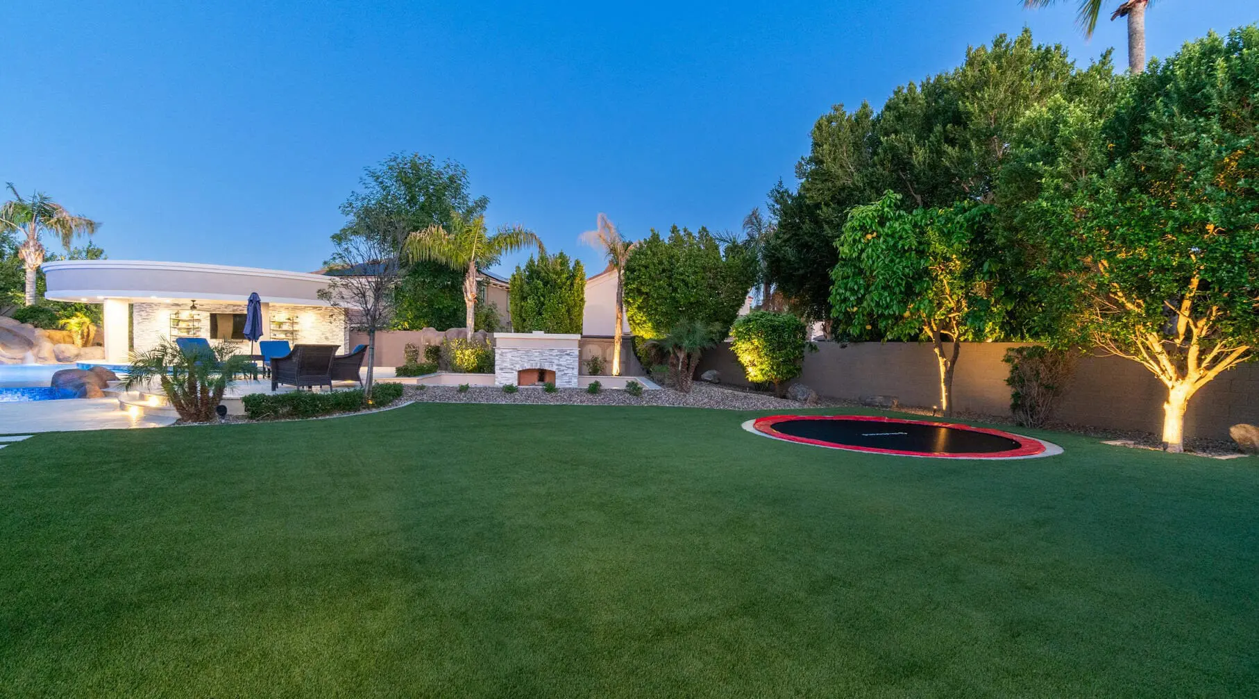 A backyard with a lush green lawn from Casa Grande Turf & Paver features an in-ground trampoline, surrounded by trees and shrubbery. In the background, there's a patio area with chairs, an outdoor fireplace, and a pool, all under a clear blue sky.