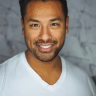 A person with short dark hair and a beard smiles warmly at the camera, wearing a white V-neck shirt. Behind them, the soft, blurred gray pattern hints at their expertise as a Casa Grande Turf & Paver specialist, enhancing spaces in Casa Grande AZ with precision and care.