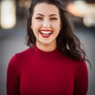 A person with long dark hair is smiling broadly, wearing a red top. The backdrop features the expertly laid surfaces of Casa Grande Turf & Paver, blending harmoniously into an outdoor setting.